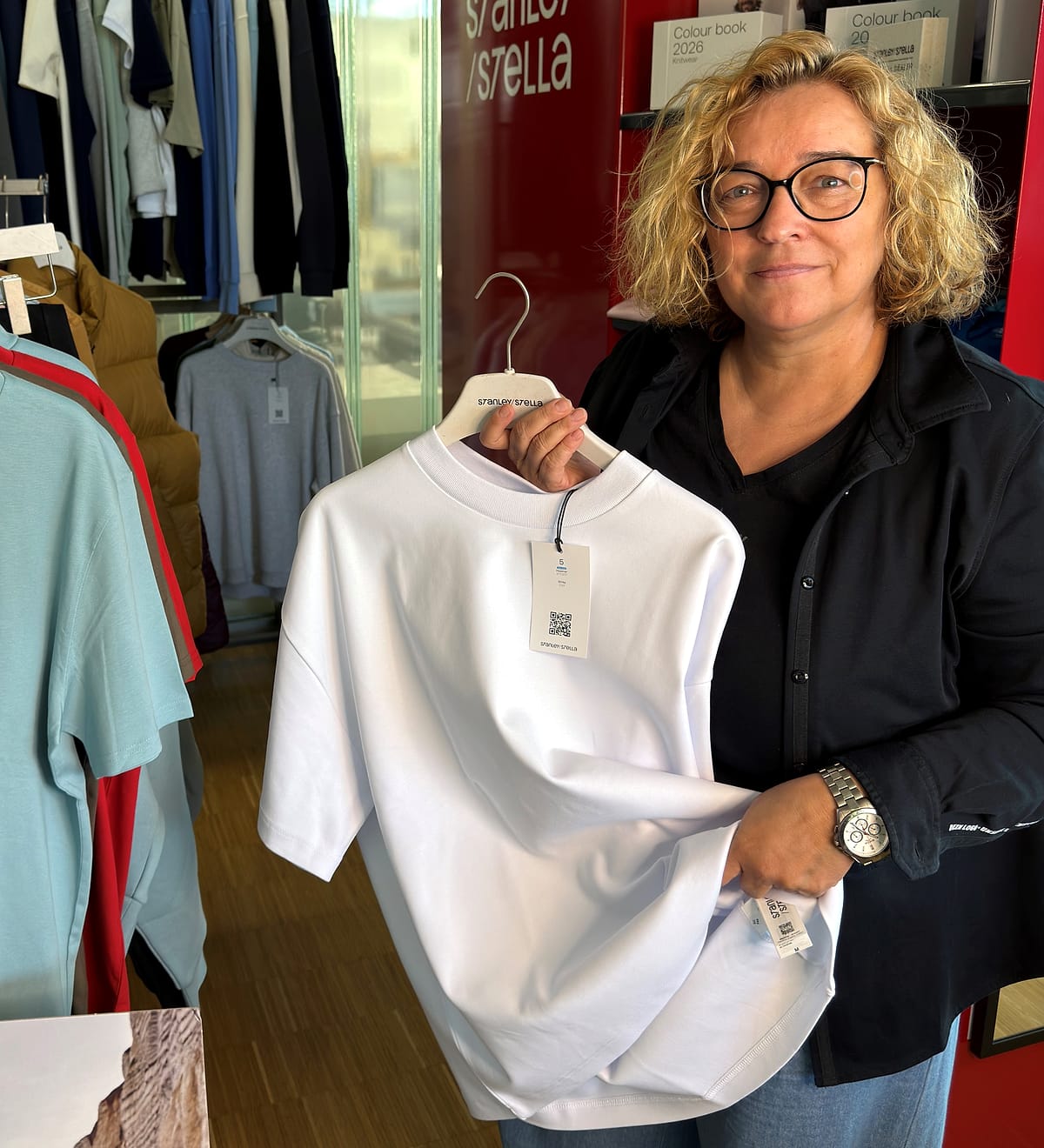 Britta Schneider hält im JobOutfit-Showroom ein weißes T-Shirt am Bügel vor die Kleiderständer