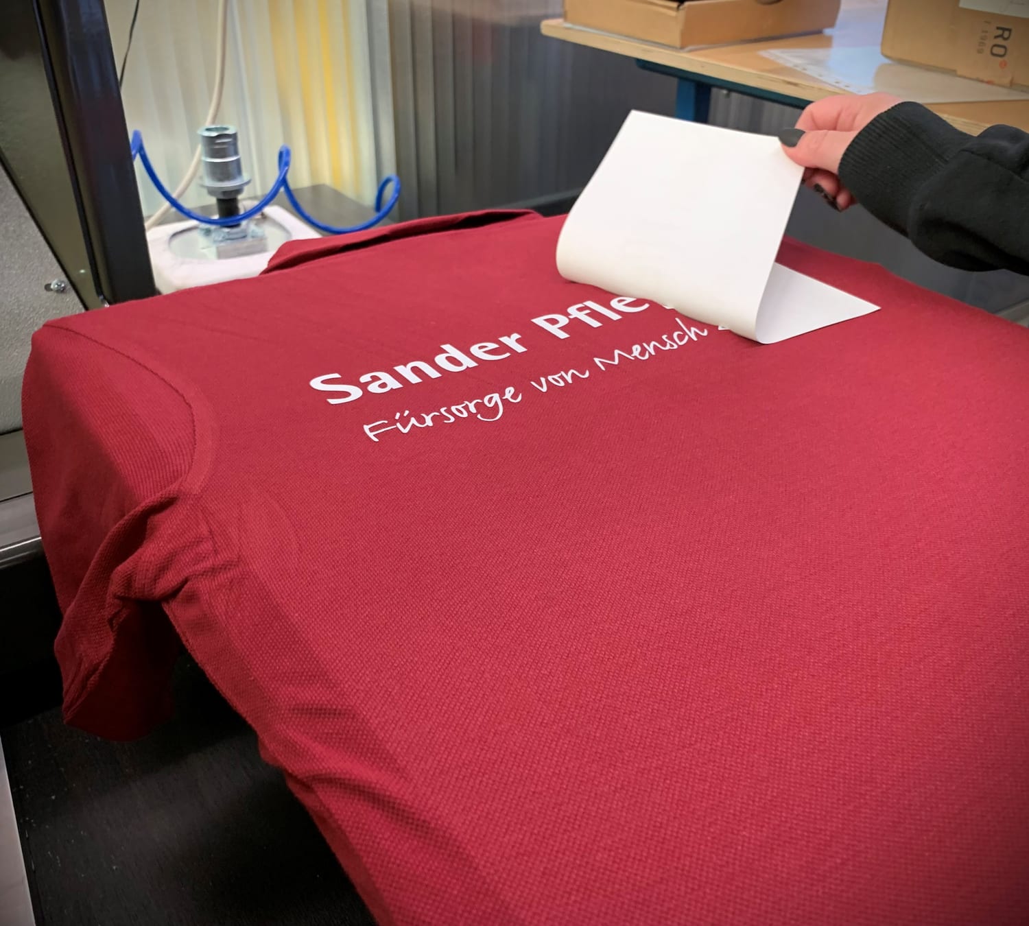 Hand zieht die Trägerfolie von einem frisch aufgepressten Logo auf einem roten Poloshirt ab